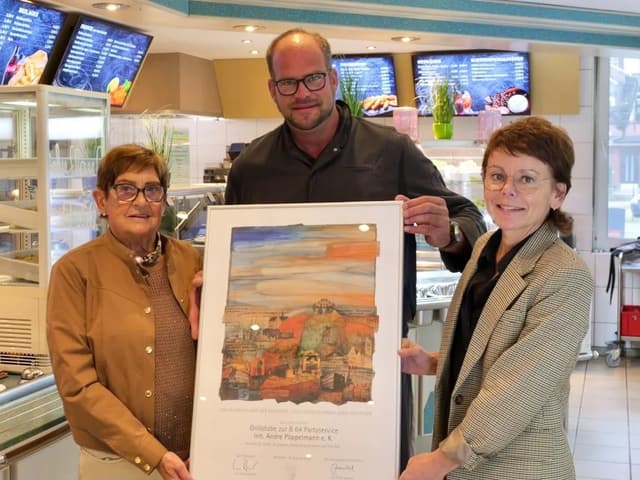 Drei Personen an der Ausgabetheke halten gemeinsam die gerahmte Jubiläumsurkunde der IHK Nord Westfalen