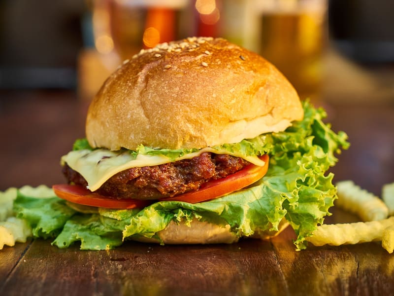 Burger mit Käse, Salat und Tomate auf einem Holzbrett, dahinter Pommes frites